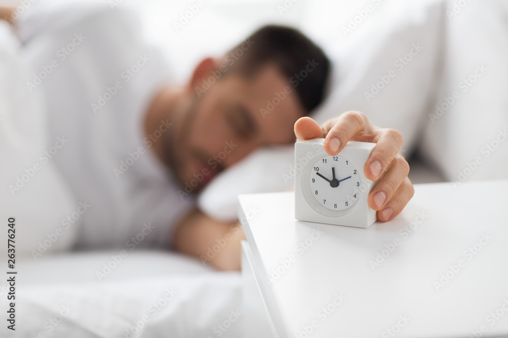 Naklejka premium morning and people concept - close up of sleepy young man in bed reaching for alarm clock on bedside table at home