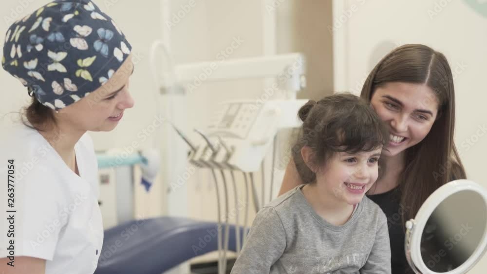 Cute little girl, her attractive mom and stomatologist are in the dental office. The girl looks at her teeth in the mirror and smiles. Cute little girl, her attractive mom and stomatologist are in the dental office. The girl looks at her teeth in the mirror and smiles.