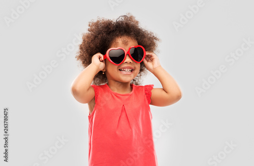 childhood, valentine's day and summer concept - happy little african american girl in heart shaped sunglasses over grey background photo