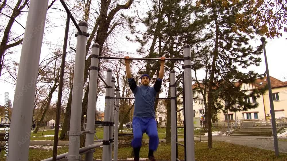 Man swings on chin up bars on exercise equipment in a public park before doing a back flip onto the ground. Guy doing Parkour stunts at a fitness station in Cluj-Napoca Park, Romania. TRACKING.