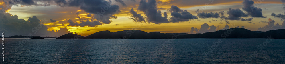 Thursday Island, Australia - February 20, 2019: Panorama of sunrise shot over Torres Strait Islands Archipelago shows dark blue clouds in yellow and red light hanging over black island hills in very d