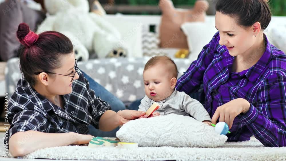Happy same sex parents playing with little cute baby lying on floor at home medium shot Happy same sex parents playing with little cute baby lying on floor at home medium shot