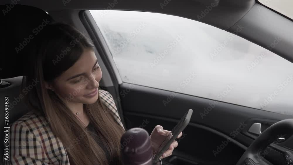 Beautiful woman driver stuck in traffic jam. She is holding a phone and a thermos in her hands. She is typing a message on the phone to somebody. Beautiful woman driver stuck in traffic jam. She is holding a phone and a thermos in her hands. She is typing a message on the phone to somebody.