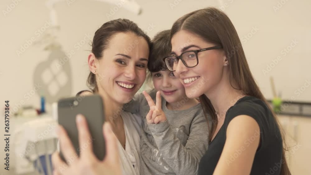 Young charming female dentist, little girl patient and her beautiful mother are making a selfie a phone after a successful appointment. They are making a funny faces. Young charming female dentist, little girl patient and her beautiful mother are making a selfie a phone after a successful appointment. They are making a funny faces.