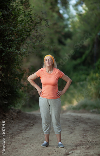 A middle-aged woman is standing on a park path with hands on her hips, tired after a jog