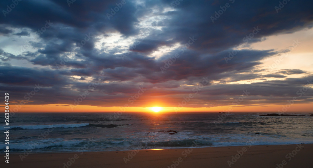 Wide angle sunset on beach in Portugal with sun on horizon and dark clouds in sky.