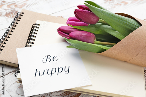 Bouquet of beautiful tulips, notebooks and card with text BE HAPPY on wooden background