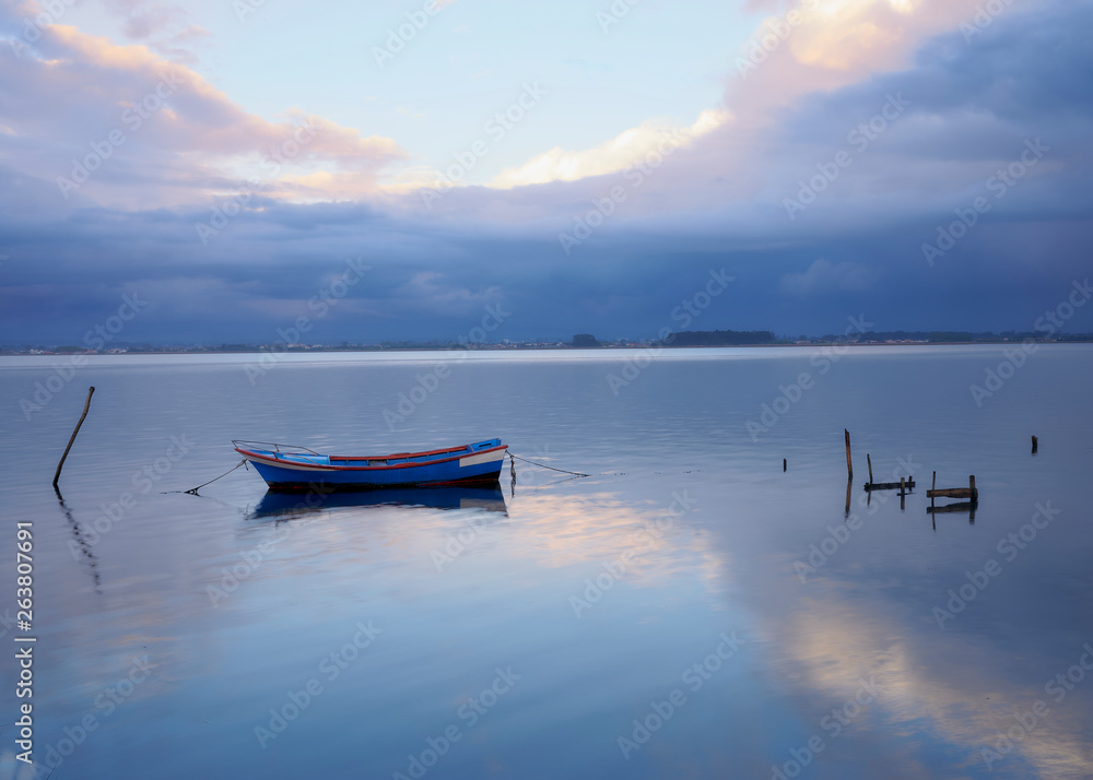 Single boat tied to a stake in the middle of the lagoon of Aveiro at Torreira. The pink and orange sunset winter clouds reflected at the lake surface. Single boat tied to a stake in the middle of the lagoon of Aveiro at Torreira. The pink and orange sunset winter clouds reflected at the lake surface.