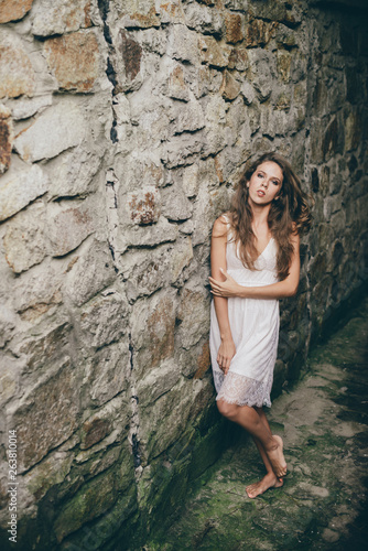 Alone beautiful girl with curly natural hair in white dress in ancient tunnel near stony mossy wall. Lonely princess in old castle. Dreamer young girl in old cool tunnel. Female beauty portrait.