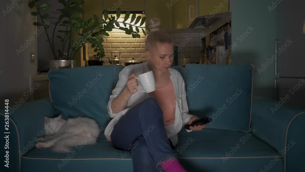 Young, attractive woman sits down with a mug on a sofa to cat and turns on the television using a remote control. 4K Young, attractive woman sits down with a mug on a sofa to cat and turns on the television using a remote control. 4K
