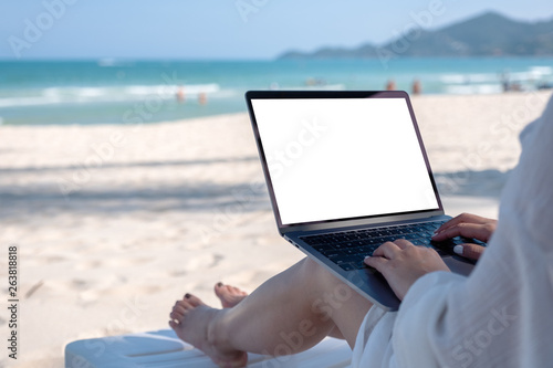 Mockup image of a woman holding and using laptop computer with blank desktop screen while laying down on beach chair on the beach
