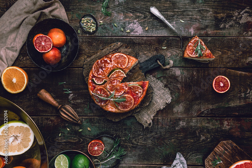 citrus upside down cake with blood oranges, lemons and limes on wooden board among fresh citrus fruit, kitchen stuff, herbs and spices on old wooden background, toned for a vintage effect