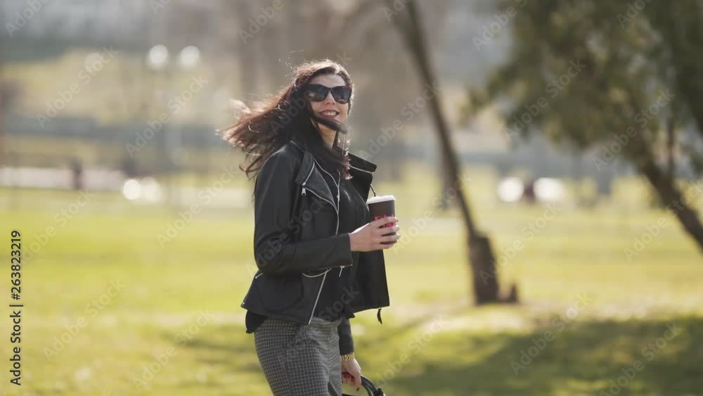 Young Caucasian Long Hair Woman in Sunglasses Walking in Public Park in Front Of Camera Holding Purse Drinking Her Coffee To Go With Straw and Smilling. Blurred Background Young Caucasian Long Hair Woman in Sunglasses Walking in Public Park in Front Of Camera Holding Purse Drinking Her Coffee To Go With Straw and Smilling. Blurred Background