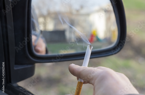 close-up, a man's hand holding a Smoking cigarette in the car window, which stands in the courtyard of a residential area overlooking a children's Playground in the rear view mirror