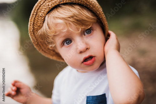 A small toddler boy standing outdoors by a river in summer.