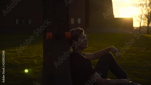 ELECTRIC LONGBOARD, 4K, SUNSET - Electric longboard leaning on a tree next to a happy young man with sunglasses that he takes off during sunset photo