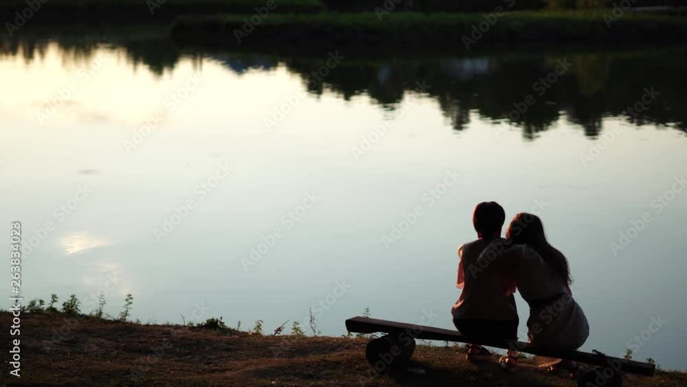 Backside view of two girls sitting on a bench by peaceful lake with reflections from trees in the golden hour Backside view of two girls sitting on a bench by peaceful lake with reflections from trees in the golden hour