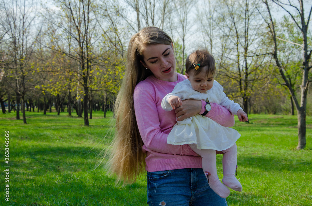 Fototapeta premium Mom with baby in bright clothes on a pink plaid on the green right. Family resting in the park on a warm day. Mom and little girl 10 months walk in the park