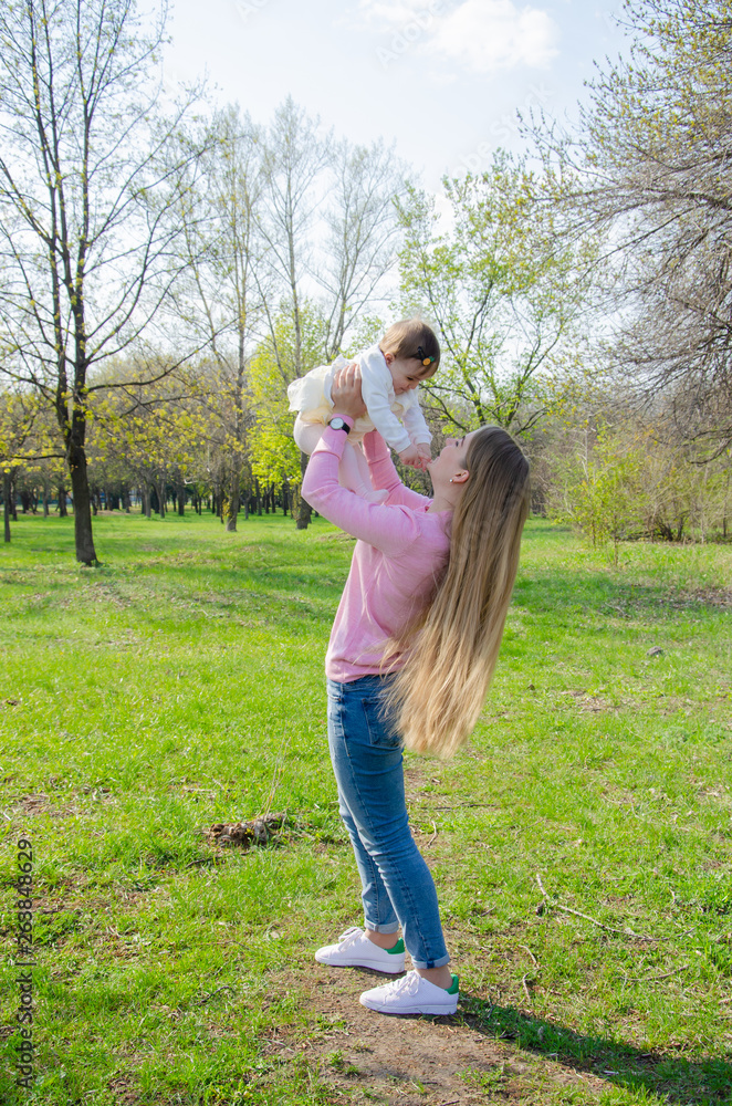 Obraz premium Mom with baby in bright clothes on a pink plaid on the green right. Family resting in the park on a warm day. Mom and little girl 10 months walk in the park