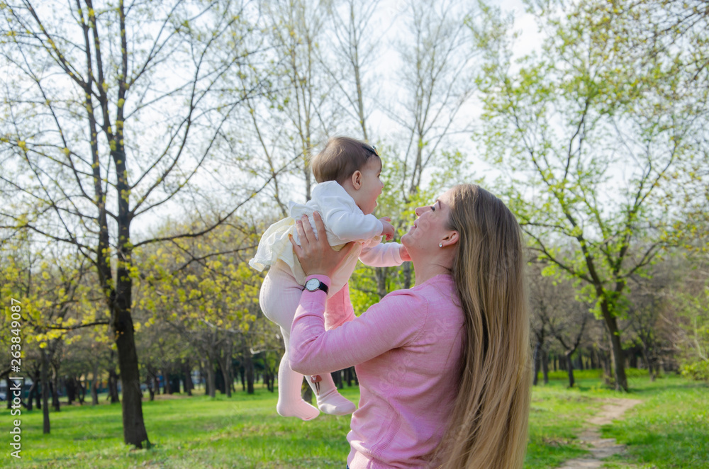 Mom with baby in bright clothes on a pink plaid on the green right. Family resting in the park on a warm day. Mom and little girl 10 months walk in the park