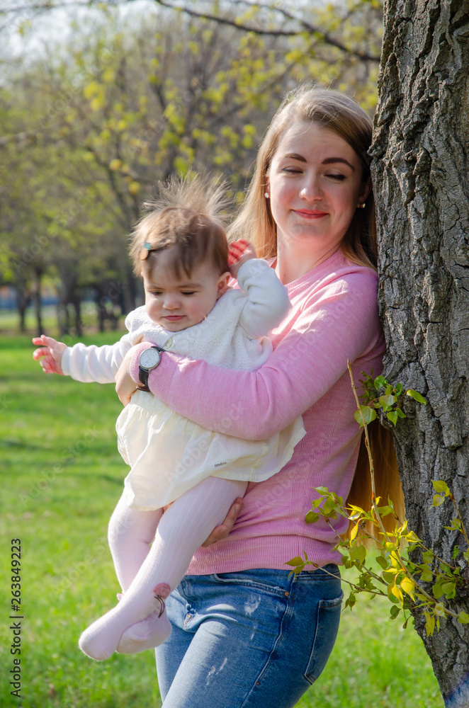 Fototapeta premium Mom with baby in bright clothes on a pink plaid on the green right. Family resting in the park on a warm day. Mom and little girl 10 months walk in the park