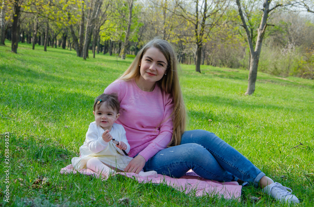 Mom with baby in bright clothes on a pink plaid on the green right. Family resting in the park on a warm day. Mom and little girl 10 months walk in the park Mom with baby in bright clothes on a pink plaid on the green right. Family resting in the park on a warm day. Mom and little girl 10 months walk in the park
