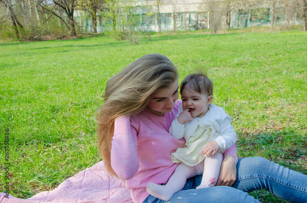 Mom with baby in bright clothes on a pink plaid on the green right. Family resting in the park on a warm day. Mom and little girl 10 months walk in the park Mom with baby in bright clothes on a pink plaid on the green right. Family resting in the park on a warm day. Mom and little girl 10 months walk in the park