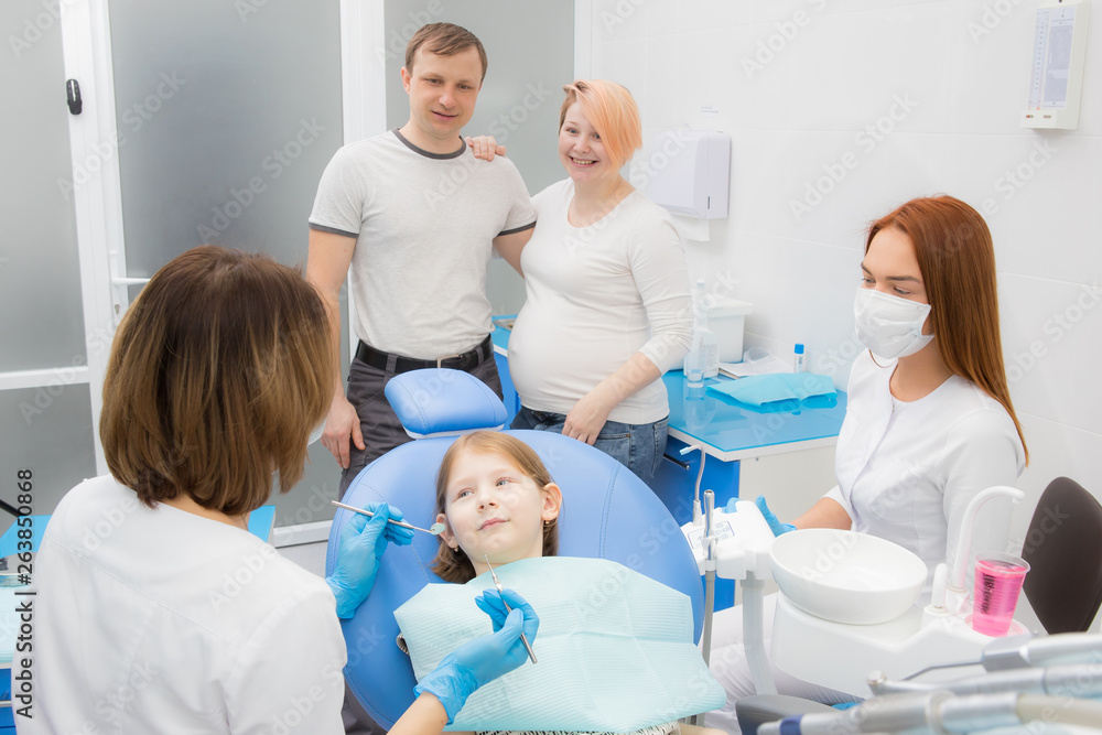 A female dentist and her assistant are treating the teeth of a little girl. Parents of the girl oversee the treatment. Mom girl is pregnant A female dentist and her assistant are treating the teeth of a little girl. Parents of the girl oversee the treatment. Mom girl is pregnant