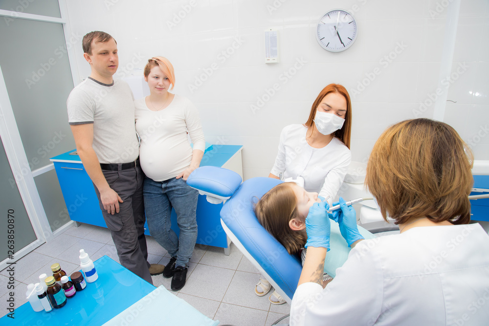 Obraz premium A female dentist and her assistant are treating the teeth of a little girl. Parents of the girl oversee the treatment. Mom girl is pregnant