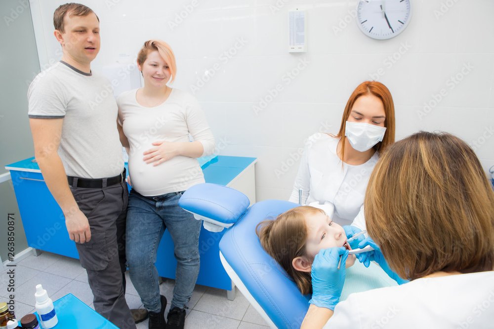 Fototapeta premium A female dentist and her assistant are treating the teeth of a little girl. Parents of the girl oversee the treatment. Mom girl is pregnant