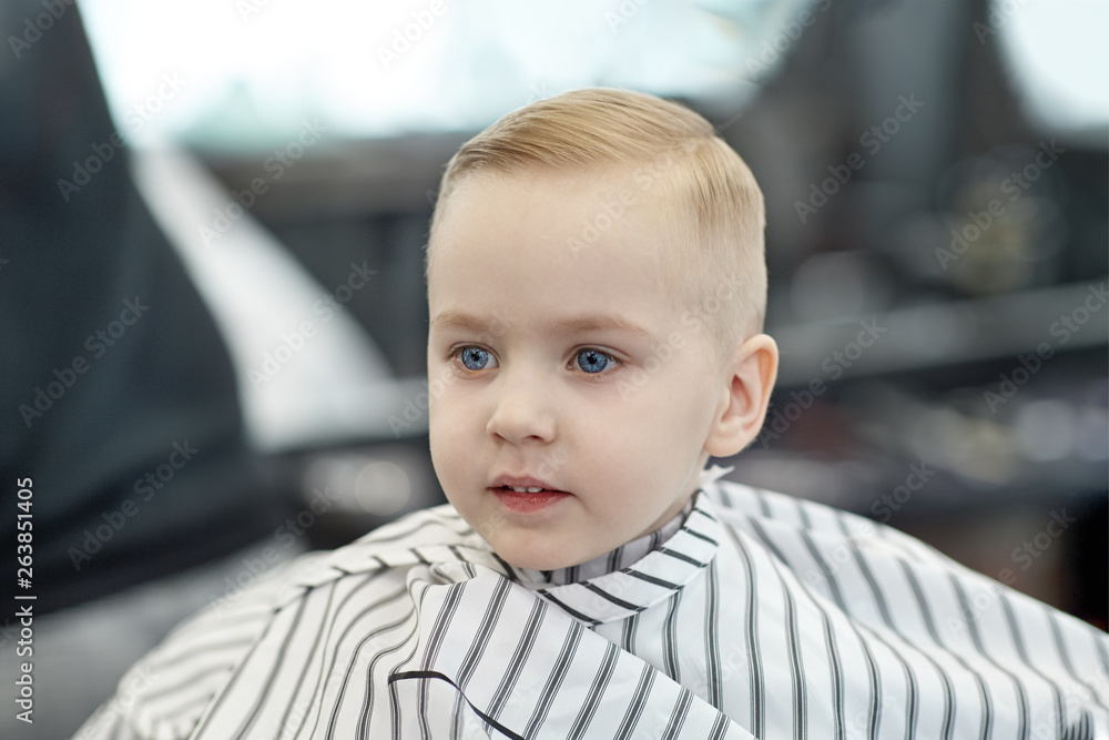 Cute blond smiling baby boy with blue eyes in a barber shop after haircut  by hairdresser. Children fashion in salon. Indoors, dark background, copy  space. Stock Photo | Adobe Stock