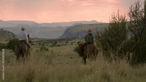 Cowboy and cowgirl ride horseback into a dramatic pink sky and mountain range backdrop, slow motion 24 fps. photo