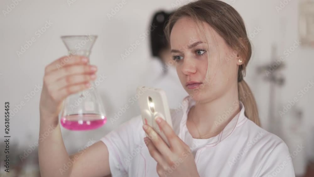 Caucasian Female Scientist in Biological Laboratory Checking Purple Liquid in Flask and Using Flashlight from Smartphone. Blurred Background Caucasian Female Scientist in Biological Laboratory Checking Purple Liquid in Flask and Using Flashlight from Smartphone. Blurred Background