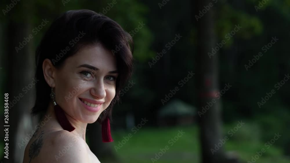 Side view of young short haired brunette with long earrings and exposed shoulders on a bokeh summer nature background turning head and smiling Side view of young short haired brunette with long earrings and exposed shoulders on a bokeh summer nature background turning head and smiling