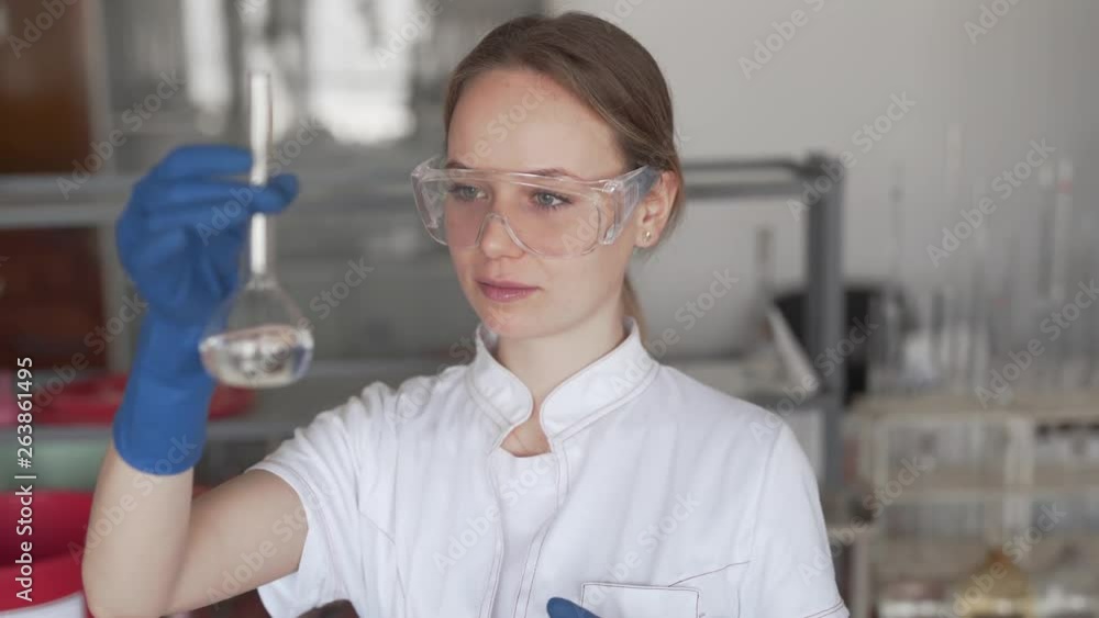 Young Female Scientist in Chemical Pesticide Laboratory Holding Volumetric Flask with Transparent Liquid Looks at Camera and Shows Sing Ok With Her Hand in Gloves. Light Camera Movement Young Female Scientist in Chemical Pesticide Laboratory Holding Volumetric Flask with Transparent Liquid Looks at Camera and Shows Sing Ok With Her Hand in Gloves. Light Camera Movement