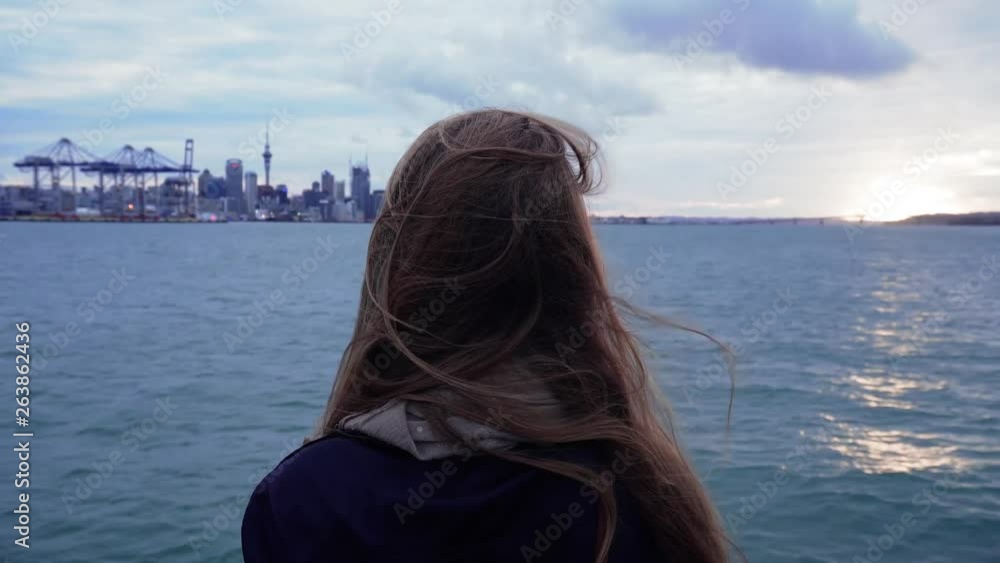 Travelling European backpacker/tourist watches the Auckland City skyline and poses in front of it while sun sets Travelling European backpacker/tourist watches the Auckland City skyline and poses in front of it while sun sets