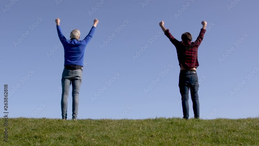 Two happy men standing up on green grass facing a blue background horizon raise their arms in victory and achievement. Shot from the back. Two happy men standing up on green grass facing a blue background horizon raise their arms in victory and achievement. Shot from the back.
