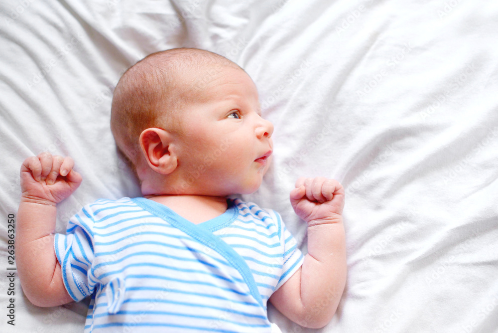 Portrait of a newborn little boy 1 months old in white blue bodysuit clothes, lying on his back in his crib on a white bedspread. Concept of health and children. Selective focus. Portrait of a newborn little boy 1 months old in white blue bodysuit clothes, lying on his back in his crib on a white bedspread. Concept of health and children. Selective focus.
