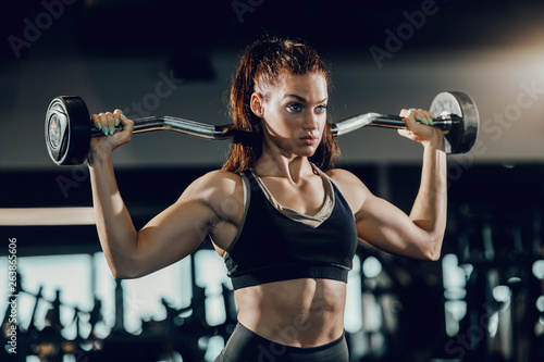 Attractive Caucasian female bodybuilder with ponytail lifting barbell while standing in gym. Train hard and do your best.