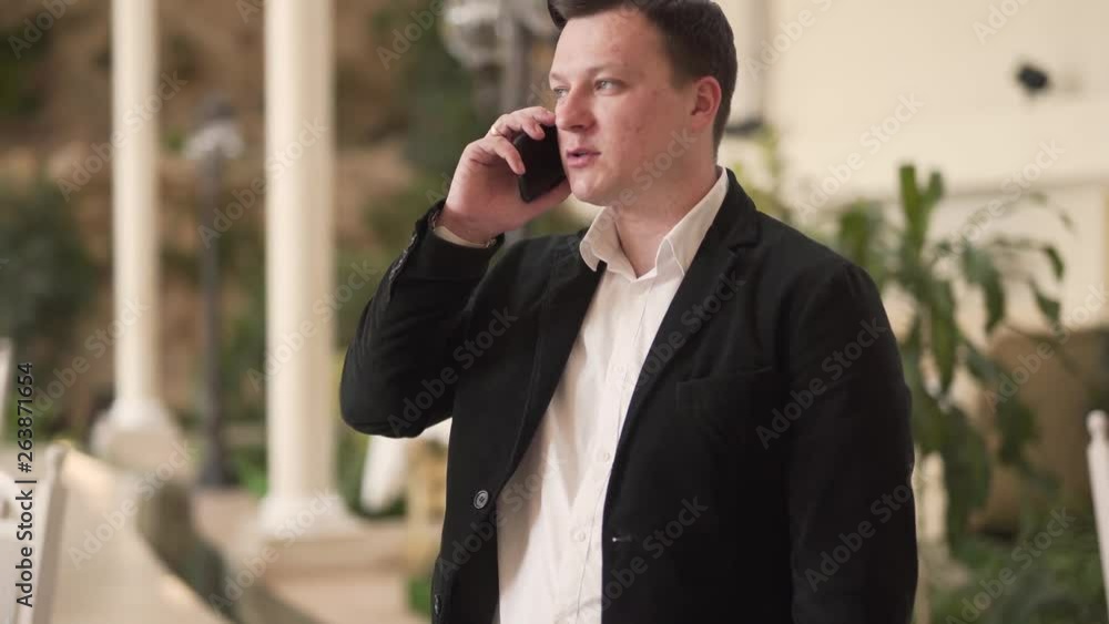 Young man in a black suit and white shirt is standing in a restaurant and talking on the phone. Man is pensive and serious. Young man in a black suit and white shirt is standing in a restaurant and talking on the phone. Man is pensive and serious.