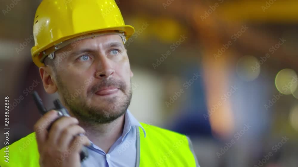 Close-up of manufacturing unit controller dressed in high-vis neon safety jacket and hardhat talking on walkie-talkie, with bright welding sparks flying in background Close-up of manufacturing unit controller dressed in high-vis neon safety jacket and hardhat talking on walkie-talkie, with bright welding sparks flying in background