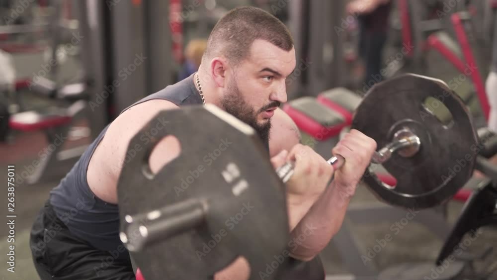 A sportsman sits on bench and lifts a barbell. He pumps lots of iron and weight in order to be fit and build more muscles. He wears a black tank-top, a beard and has a chain with pendant on his neck. A sportsman sits on bench and lifts a barbell. He pumps lots of iron and weight in order to be fit and build more muscles. He wears a black tank-top, a beard and has a chain with pendant on his neck.