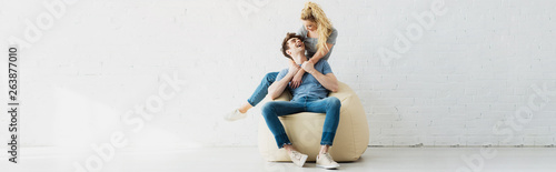 panoramic shot of cheerful blonde girl hugging happy boyfriend sitting on beige bean bag chair at home