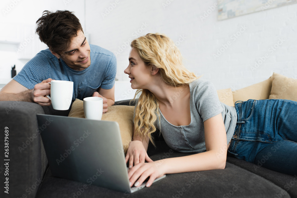 happy and handsome man holding cups near blonde girl using laptop