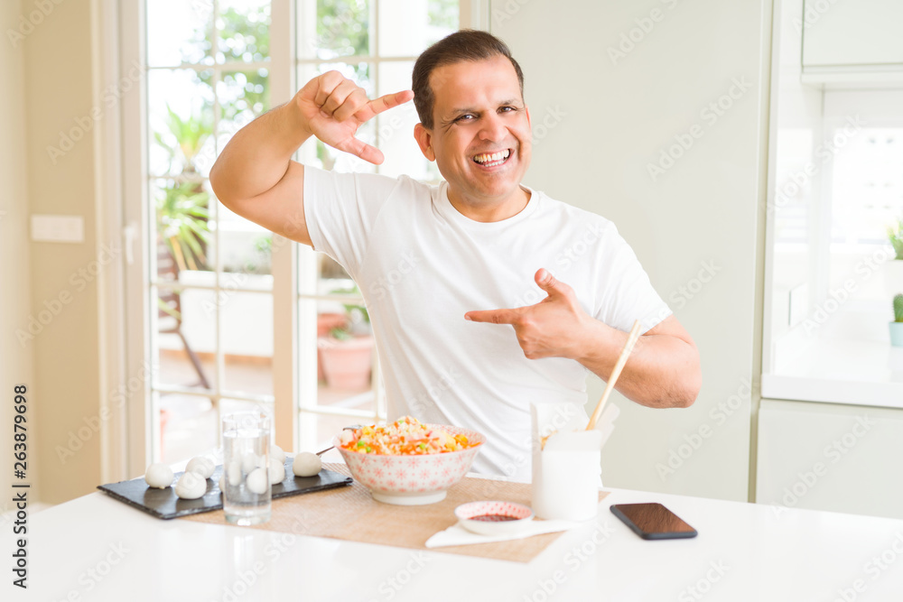 Middle age man eating asian food with chopsticks at home smiling making frame with hands and fingers with happy face. Creativity and photography concept.