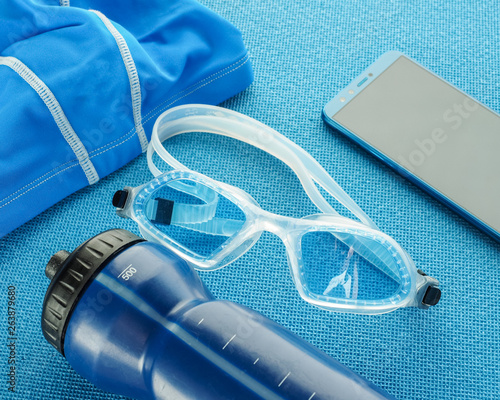 Flat lay composition with Goggles, swimming cap, smartphone, earphones, fitness tracker and bottle of water equipment for swimming pool on blue background. top view. Swimming set.