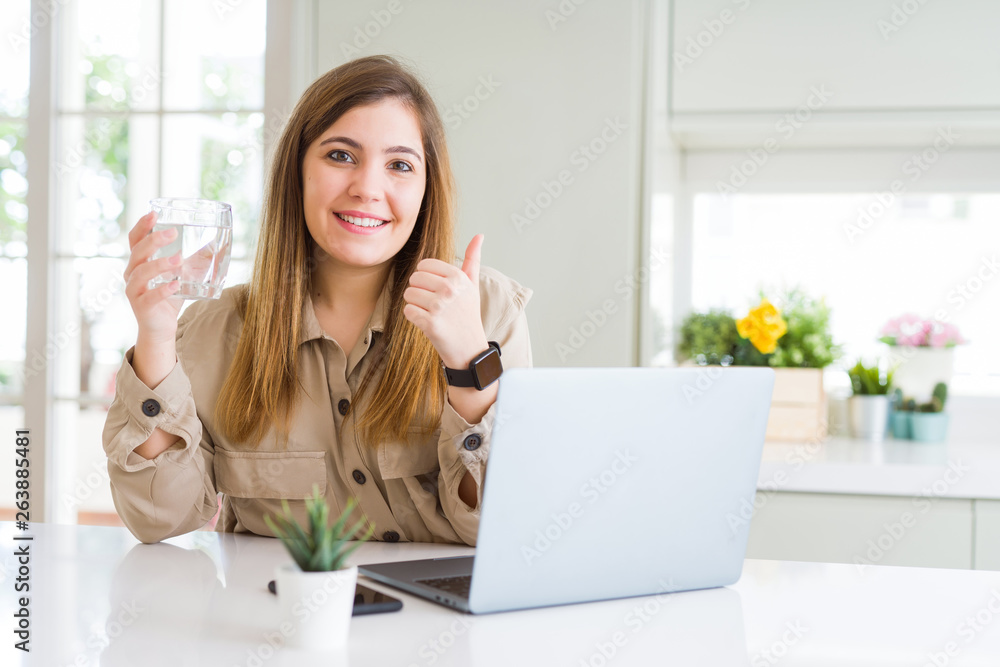 Beautiful young woman working with computer takes a break to drink glass of water happy with big smile doing ok sign, thumb up with fingers, excellent sign Beautiful young woman working with computer takes a break to drink glass of water happy with big smile doing ok sign, thumb up with fingers, excellent sign