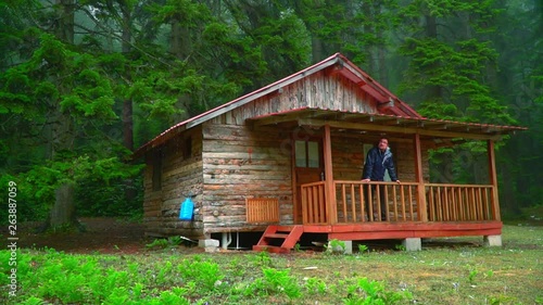 camper man watching view from balcony of wooden house while raining in forest