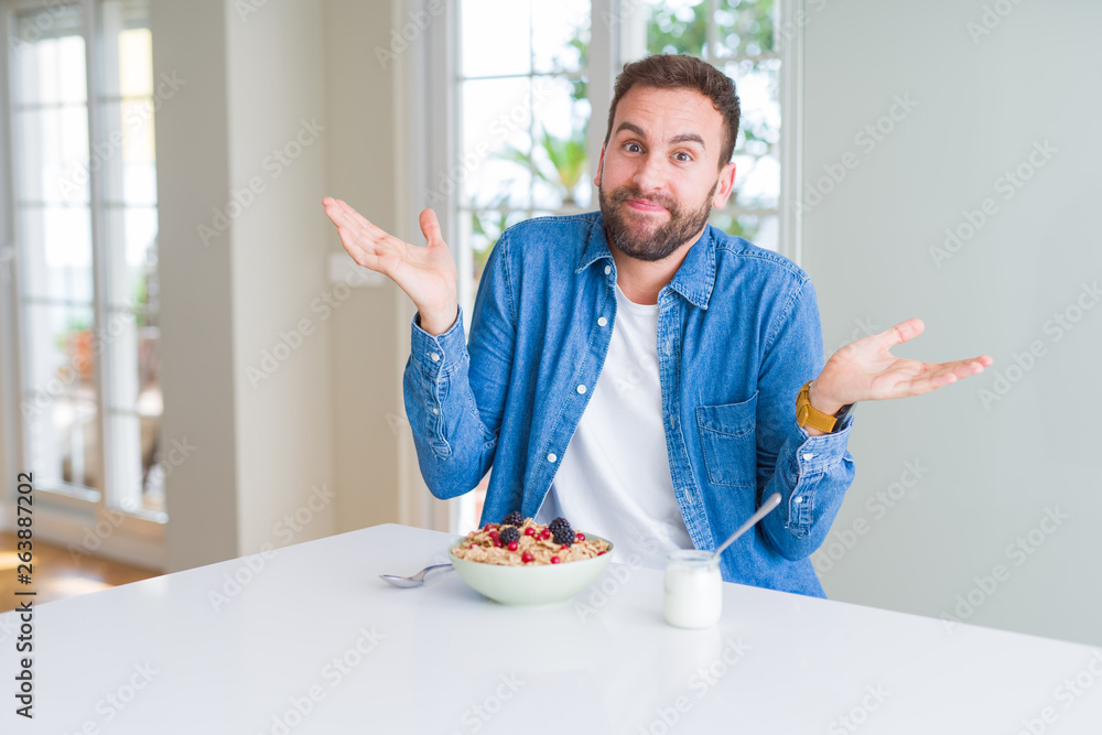 Handsome man eating cereals for breakfast at home clueless and confused expression with arms and hands raised. Doubt concept. Handsome man eating cereals for breakfast at home clueless and confused expression with arms and hands raised. Doubt concept.