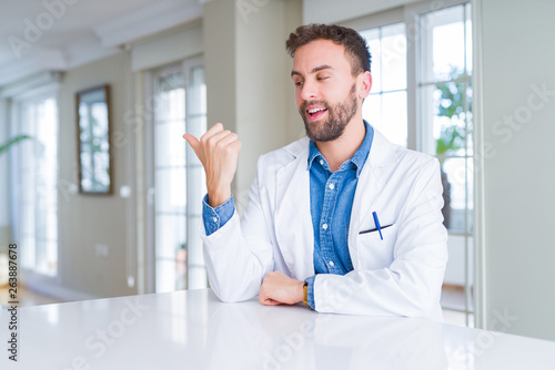 Handsome doctor man wearing medical coat at the clinic smiling with happy face looking and pointing to the side with thumb up.